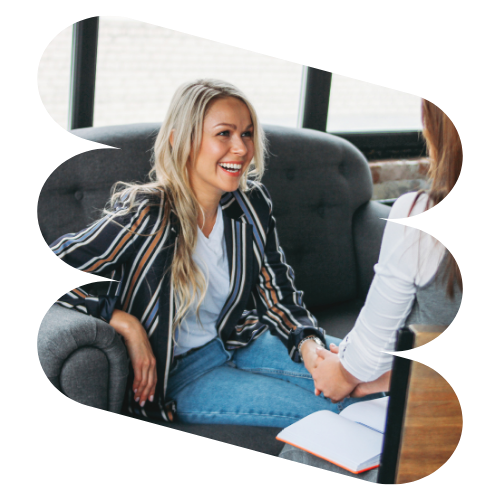A woman with long blonde hair, wearing a striped blazer and jeans, sits on a gray sofa in a well-lit room with large windows. She smiles while discussing ideas about resilience with another person who holds a notebook.