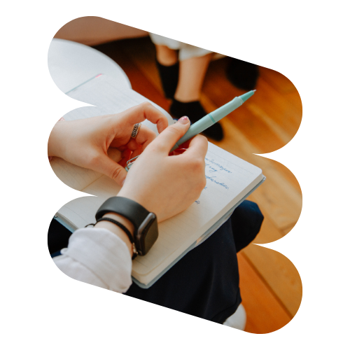 A person focused on mental health treatment in Chicago holds a turquoise pen, jotting down thoughts in a notebook while sitting on a wooden floor. Adorned with a watch and rubber band, only their hands and part of the notebook come into view.