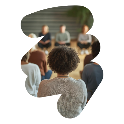 In a softly lit room with wooden floors and plants, a group sits in a circle, engaged in a communal discussion about resilience. The focus is on an individual with curly hair, seen from behind, as they share insights and foster connection in this warm gathering.