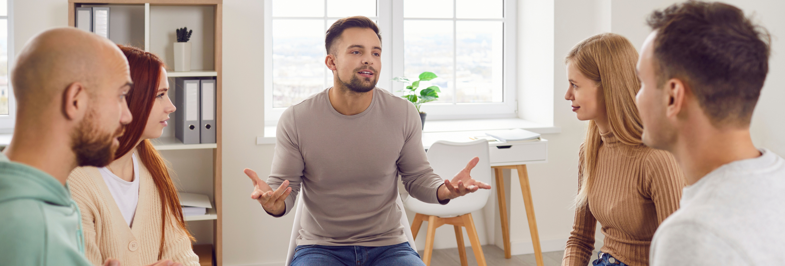 In a bright room in Chicago, a group of five sits in a circle discussing depression treatment. The scene includes two women and three men, with shelves and a window view providing the backdrop to their conversation.