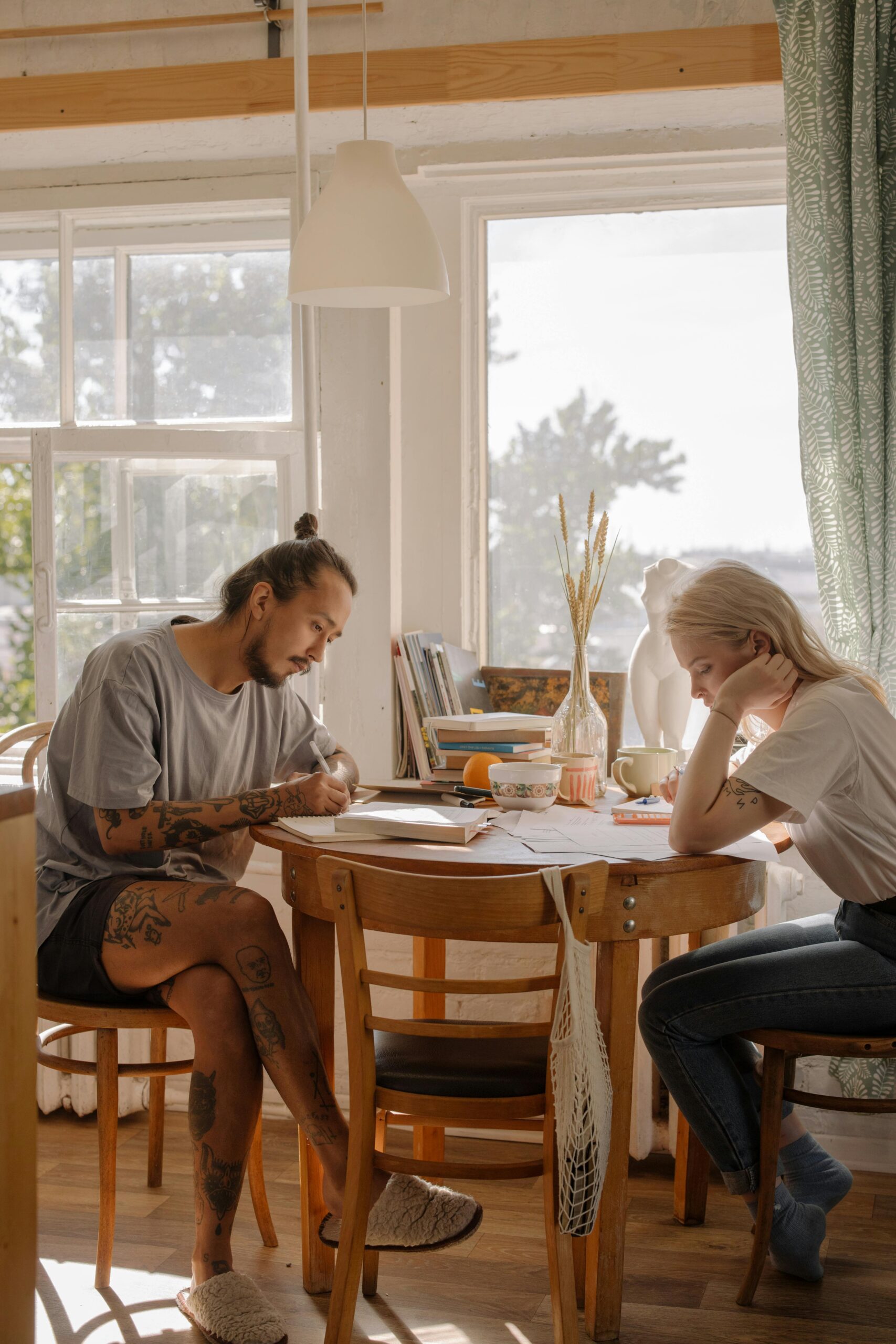 Two people sit at a wooden table in a sunlit room, each focused on writing or reading—an inviting scene that reflects the calm and focus often encouraged in adult mental health treatment. Books, fruit, and mugs add to the cozy, studious atmosphere.